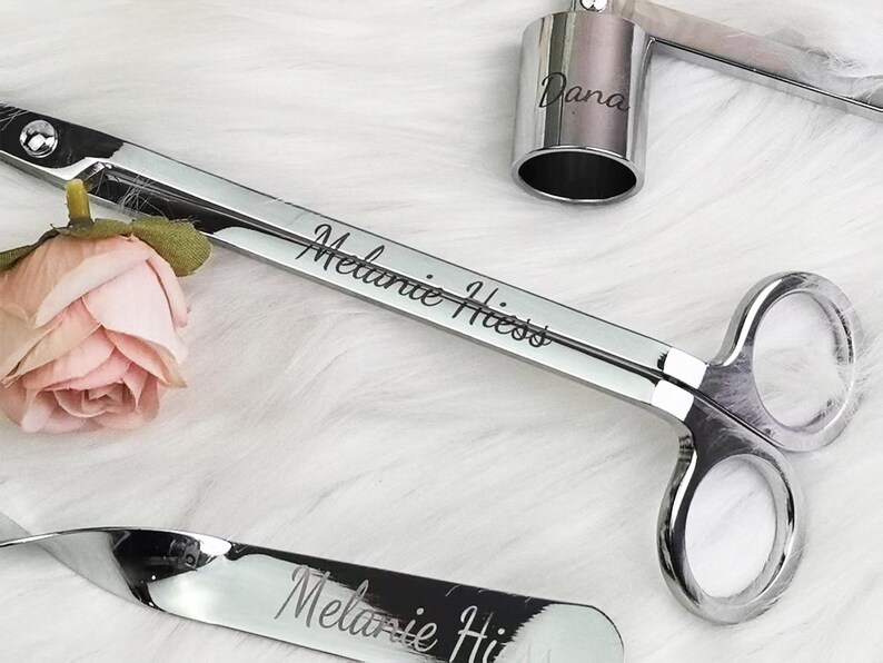Op de afbeelding: Een set zilveren kaarsaccessoires, waaronder een lonttrimmer, een snuffer en een kaarshouder. De lonttrimmer en snuffer zijn gegraveerd met de naam "Melanie Hiess". Een kleine roze roos staat naast de trimmer.