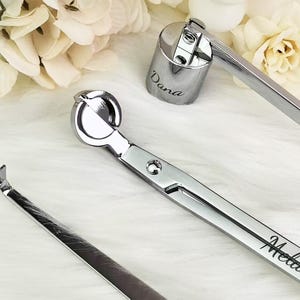 Op de afbeelding: Zilveren kaarsenaccessoireset, inclusief een lonttrimmer, een lontdompelaar en een kaarsensnuffer. De lonttrimmer en snuffer zijn gegraveerd met namen. Gepresenteerd op een witte, zachte ondergrond.