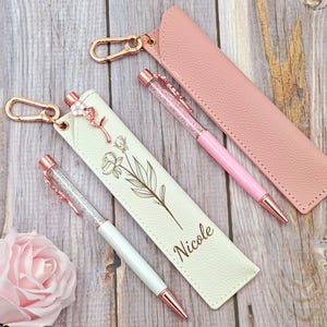 May include: A selection of pens and pen cases in rose gold and pink hues. One pen case is white with a floral design and the name "Nicole". The pens feature crystal-filled barrels and rose gold detailing.