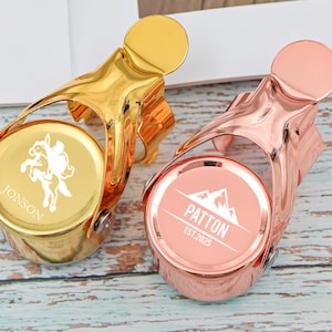 May include: Two metal champagne bottle stoppers. One is gold with a rider graphic and the name "Jonson". The other is rose gold with a mountain graphic and the name "Patton" and the year "EST. 2025".