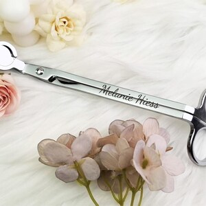 Op de afbeelding: Een zilveren kaarsenpit trimmer met de naam "Melanie Hess" gegraveerd op het handvat. De trimmer heeft een cirkelvormig mes en schaarachtige handvatten. De afbeelding bevat roze en witte bloemen.