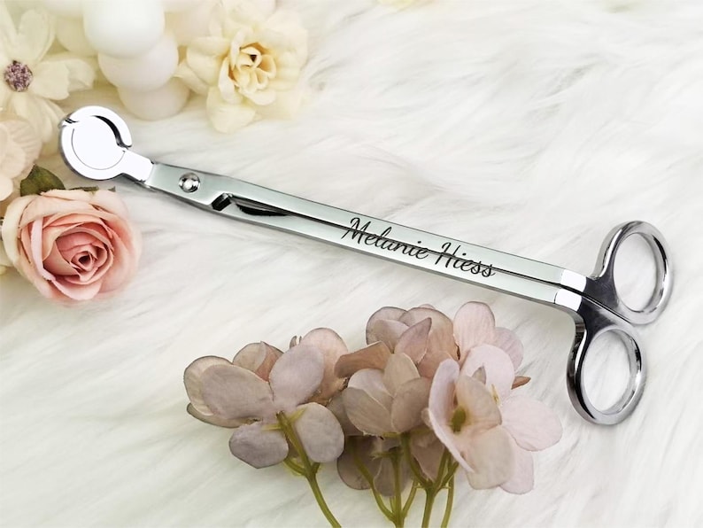 Op de afbeelding: Een zilveren kaarsenpit trimmer met de naam "Melanie Hess" gegraveerd op het handvat. De trimmer heeft een cirkelvormig mes en schaarachtige handvatten. De afbeelding bevat roze en witte bloemen.