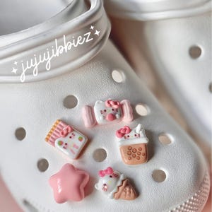 Peut inclure: Sabots blancs avec des breloques de chaussures sur le thème Hello Kitty roses et blanches. Les breloques comprennent une étoile, un Hello Kitty tenant une sucette glacée, un Hello Kitty tenant une tasse de bubble tea, un Hello Kitty tenant une boîte de bonbons et un Hello Kitty tenant une canne en sucre.