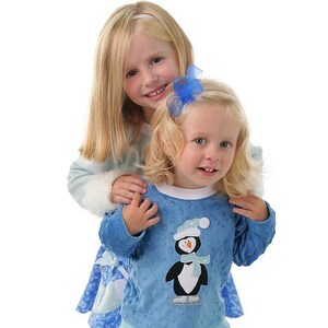 May include: Two young girls wearing blue and white winter outfits. The girl in the front is wearing a blue long-sleeved shirt with a penguin graphic and a white trim. The girl in the back is wearing a blue and white dress with a white fur trim. Both girls have blonde hair.