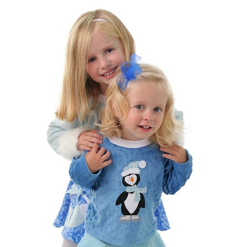 May include: Two young girls wearing blue and white winter outfits. The girl in the front is wearing a blue long-sleeved shirt with a penguin graphic and a white trim. The girl in the back is wearing a blue and white dress with a white fur trim. Both girls have blonde hair.
