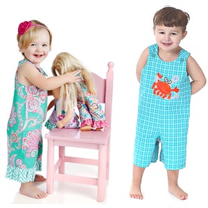 May include: Two children wearing summer outfits. A girl in a green and pink floral dress is holding a doll. A boy in blue and white checkered overalls with a red crab applique is standing with his hands in his pockets.