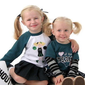 May include: Two young girls wearing cheerleading outfits. One girl is wearing a green and white shirt with a cheerleader graphic and a black skirt. The other girl is wearing a teal shirt with a heart and the text "Heart of the Team" and a black and white polka dot skirt. Both girls have their hair in pigtails.