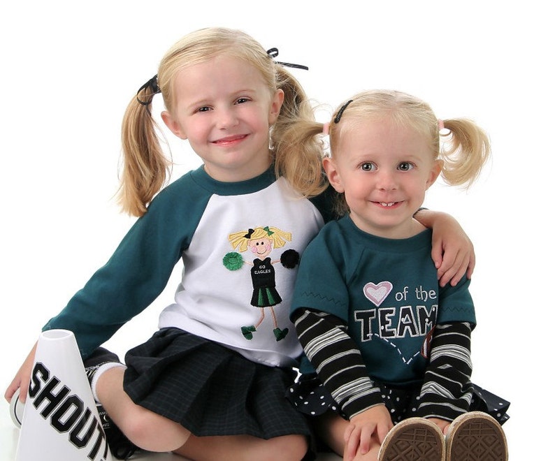 May include: Two young girls wearing cheerleading outfits. One girl is wearing a green and white shirt with a cheerleader graphic and a black skirt. The other girl is wearing a teal shirt with a heart and the text "Heart of the Team" and a black and white polka dot skirt. Both girls have their hair in pigtails.