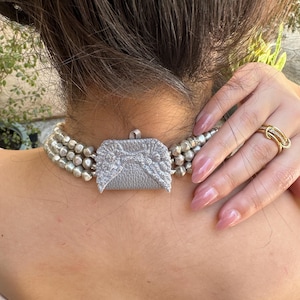 May include: A multi-strand necklace featuring metallic silver beads and a central rectangular pendant with a textured silver finish and glittery angel wing embellishments. A gold-toned ring with a multi-band design is also visible.