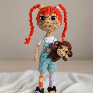 May include: A crocheted doll with bright orange hair in two long braids, wearing a white shirt, light blue overalls with colorful buttons, and mismatched striped socks. The doll has black shoes with purple and yellow laces and holds a small brown monkey.