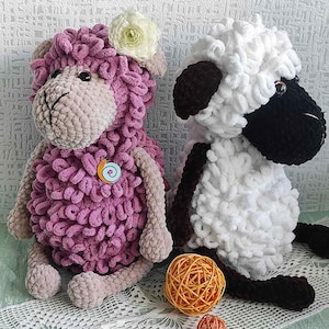 May include: Two handmade crochet sheep dolls. One is lavender with a beige face and limbs, and a white flower on its head. The other is white with black accents. Two small yarn balls in orange and brown are in front of the sheep.