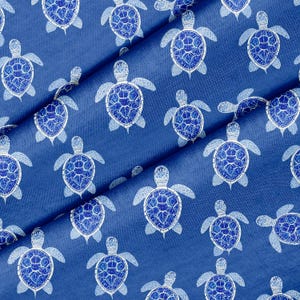 May include: A blue fabric featuring a repeating pattern of sea turtles in white and blue. The turtles have detailed shell patterns and are evenly distributed. The fabric is folded, revealing its texture and depth.