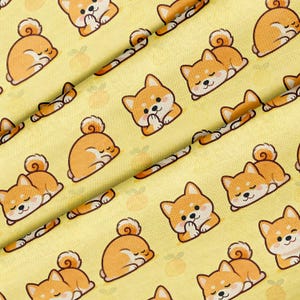 Peut inclure: Un tissu jaune avec un motif répété de chiens Shiba Inu de dessin animé en orange et blanc. Certains chiens dorment, d'autres sont assis, les pattes jointes. Le fond comprend de petites illustrations de fruits oranges.