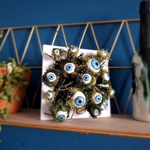 May include: A white square canvas with a cluster of blue and white eyeballs embedded in green moss. The eyeballs are arranged in a random pattern and appear to be staring out at the viewer.