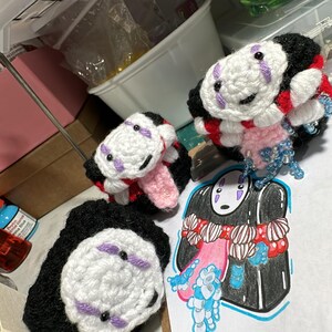 May include: Handmade crochet No-Face plushies and a No-Face illustration. The plushies feature a white face with purple accents, black bodies, and red and pink details. The illustration is a colorful drawing of the character.