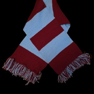 May include: A knitted scarf with alternating maroon and light blue stripes. The scarf has fringed ends with white and light blue threads. The scarf is displayed against a black background.