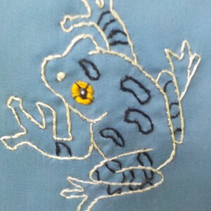 May include: A blue fabric with a white embroidered frog with a yellow eye. The frog is in a sitting position with its legs outstretched.