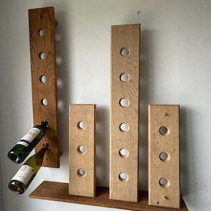 Puede incluir: Un estante para vino de pared hecho de madera marrón claro con cinco filas de recortes circulares para sostener botellas de vino. Dos botellas de vino se muestran en el estante. El estante está montado en una pared blanca.