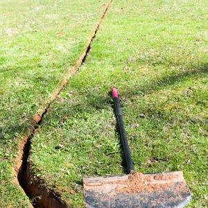 May include: A black garden tool with a red handle is used to dig a trench in a grassy lawn. The trench is about 10 feet long and 6 inches wide.