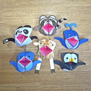 Può includere: Sei giocattoli di origami di animali di carta colorati con la bocca aperta. Gli animali sono un panda, un procione, una girafa, una balena, uno squalo e un pinguino.