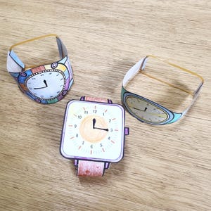 Può includere: Tre artigianato di orologi di carta con elastici. Gli orologi hanno quadranti e lancette di diversi colori. Un orologio ha un quadrante quadrato con numeri arancioni e un centro giallo. Un altro orologio ha un quadrante rotondo con numeri colorati e un centro bianco. Il terzo orologio ha un quadrante rotondo con numeri blu e un centro giallo.