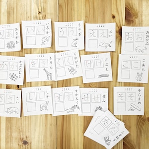 Op de afbeelding: Een set van 12 flashcards met Japanse karakters en illustraties van dieren. Elke kaart heeft een ander karakter en dier, zoals een giraffe, een kever, een dolfijn en een koala.