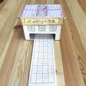 May include: A paper model of a ticket booth with the text "バッチーハウス" on the front. A paper ticket with handwritten numbers is coming out of the booth.