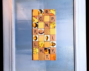 May include: A framed artwork featuring a vertical arrangement of letter tiles spelling out the word "FAMILY". The tiles are decorated with various images of fruits, flowers, and other nature-inspired designs. The background is a light blue canvas.