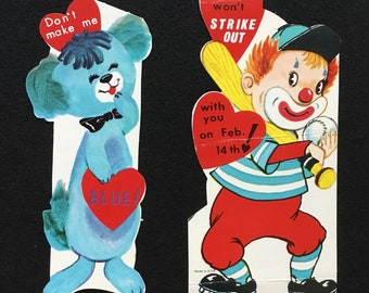 May include: Two vintage Valentine's Day cards. The first card features a blue cartoon dog holding a red heart with the word "BLUE!" on it. The second card features a cartoon clown holding a baseball bat and a baseball. The clown is wearing a red and white striped shirt and red pants. The card says "I hope I won't strike out with you on Feb. 14th."
