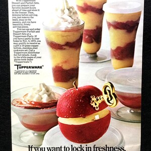 Könnte beinhalten: Eine alte Tupperware-Werbung mit hohen Parfaitgläsern, gefüllt mit gelben und roten Schichten und Schlagsahne. Ein roter Apfel mit einem goldenen Schlüsselmechanismus obenauf steht auf einer weißen Untertasse im Vordergrund.
