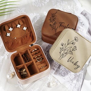 Może przedstawiać: Dwa małe, kwadratowe pudełka na biżuterię ze skóry w kolorze brązowym z kwiatowymi wzorami i spersonalizowanymi nazwami. Pudełko po lewej stronie jest otwarte i pokazuje przegródki na pierścionki, kolczyki i inne ozdoby. Pudełko po prawej stronie jest zamknięte i ma wygrawerowane imię "Vicky".