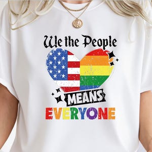 May include: White t-shirt featuring a heart design split into the American flag and a rainbow. The text reads "We the People MEANS EVERYONE" in black and rainbow colors. The shirt is a casual wear item.