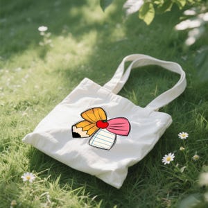 May include: A white canvas tote bag with a colorful graphic of a pencil, apple, and pink bow. The bag is sitting on a bed of green grass with small white flowers. The tote bag has two white handles.