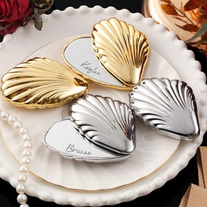 May include: Gold and silver seashell-shaped place card holders on a white plate with a gold rim. The place cards have names written on them. A pearl necklace and red roses are in the background.