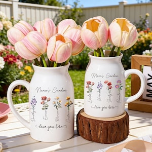 Puede incluir: Dos jarrones de cerámica blanca con asas, cada uno con tulipanes rosas y amarillos. Uno de los jarrones dice "Nana's Garden" y el otro "Mom's Garden". Ambos jarrones tienen detalles florales y nombres, con la frase "love you here."