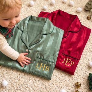 May include: Two button-down pajama shirts with long sleeves and collars. One shirt is sage green with the embroidered gold letters LHB on the pocket. The other shirt is deep red with the embroidered gold letters JEP on the pocket.
