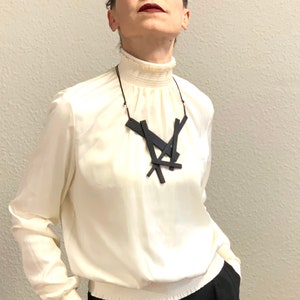 May include: A cream-colored blouse with a high, ruffled collar and long sleeves, paired with black trousers. A black geometric pendant necklace hangs from a thin cord. The outfit is simple and elegant.
