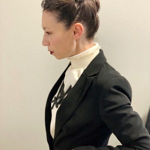 May include: A black blazer with a white blouse featuring a ruffled collar. A black necklace with a geometric design is visible. The person's hair is styled in a bun. The overall look is professional and stylish.