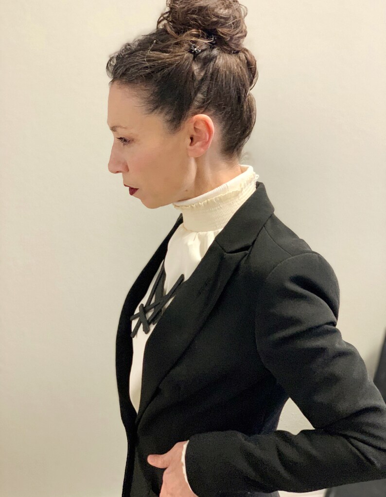 May include: A black blazer with a white blouse featuring a ruffled collar. A black necklace with a geometric design is visible. The person's hair is styled in a bun. The overall look is professional and stylish.