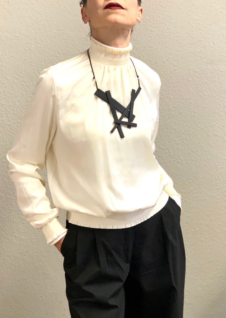 May include: A cream-colored blouse with a high, ruffled collar and long sleeves, paired with black trousers. A black geometric pendant necklace hangs from a thin cord. The outfit is simple and elegant.