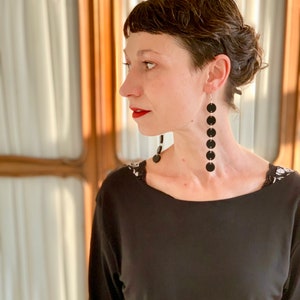 May include: A woman wearing long black earrings with five black circular pendants. She is wearing a black long-sleeved top with a scoop neckline. The background features a window with white curtains.