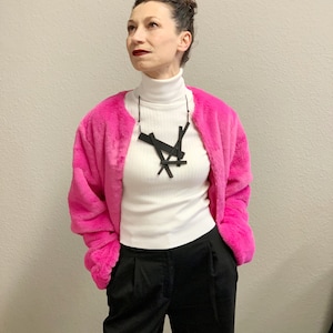 May include: A woman wearing a bright pink faux fur jacket over a white turtleneck and black pants. A black geometric pendant necklace hangs from a thin cord. The outfit is a mix of textures and colors.