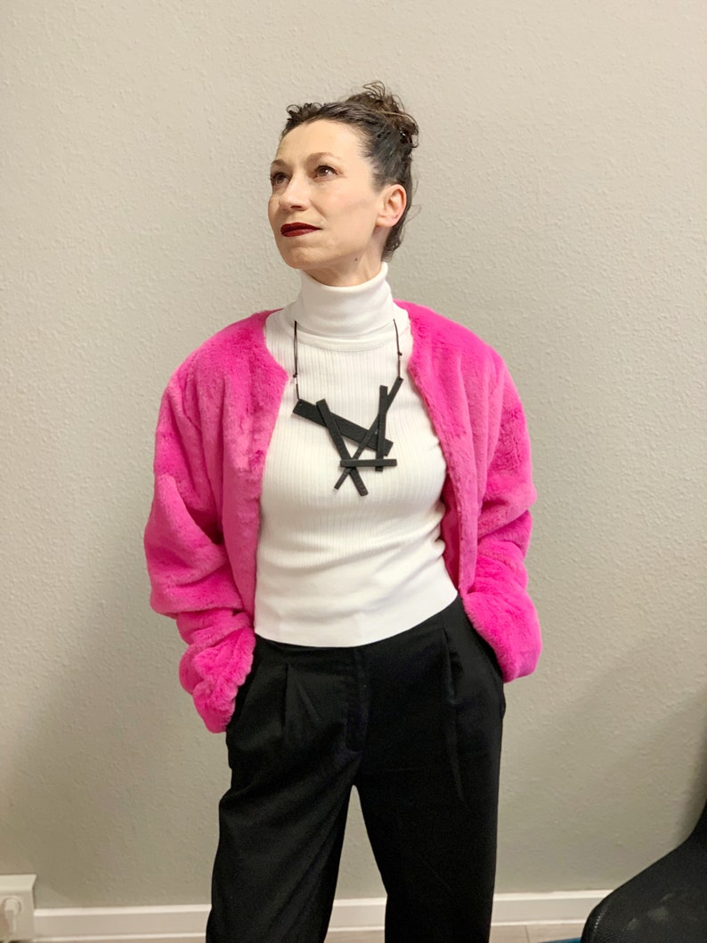 May include: A woman wearing a bright pink faux fur jacket over a white turtleneck and black pants. A black geometric pendant necklace hangs from a thin cord. The outfit is a mix of textures and colors.