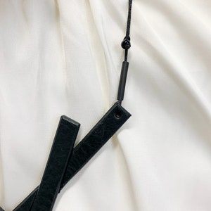 May include: Black leather belt with a black cord and a small black bead. The belt has two rectangular leather pieces that cross each other. The background is a white fabric.