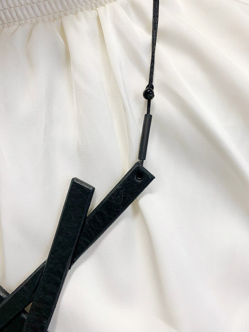 May include: Black leather belt with a black cord and a small black bead. The belt has two rectangular leather pieces that cross each other. The background is a white fabric.