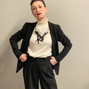 May include: A woman wearing a black blazer, black trousers, and a cream-colored blouse with a high ruffled collar. A black geometric pendant necklace is the focal point. The outfit is a classic, professional ensemble.