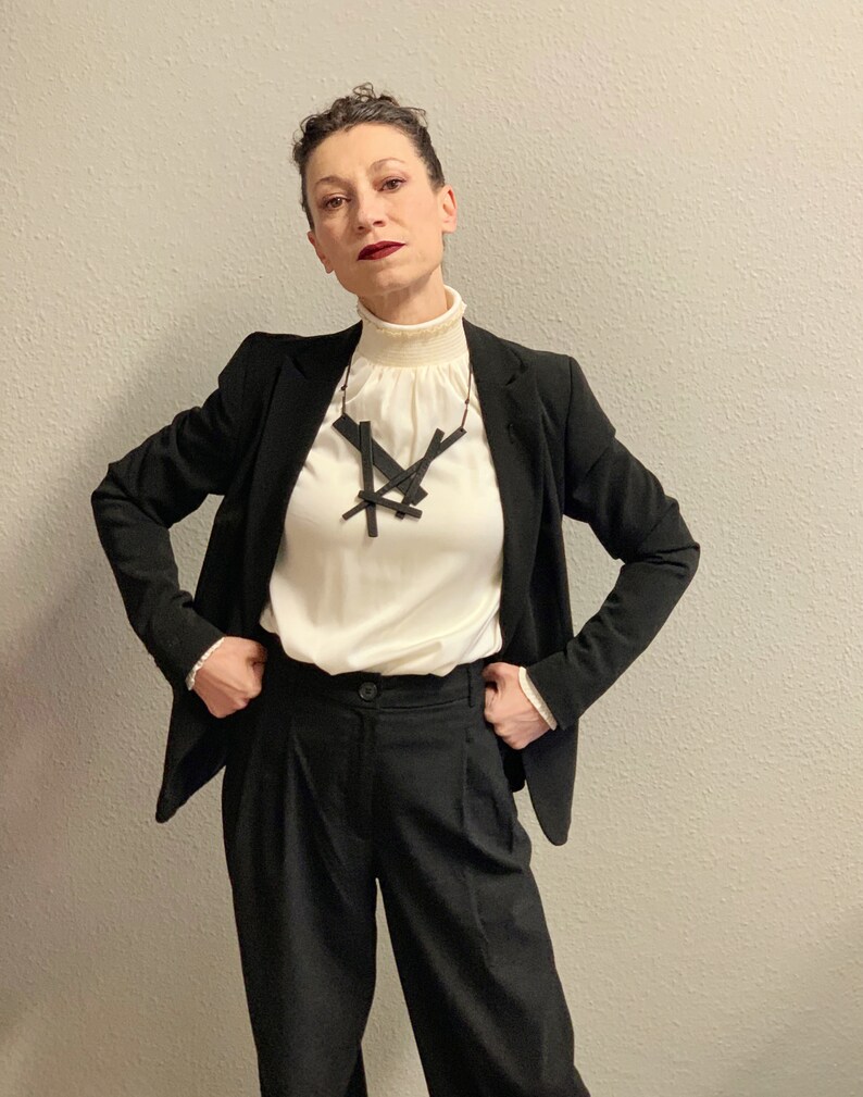 May include: A woman wearing a black blazer, black trousers, and a cream-colored blouse with a high ruffled collar. A black geometric pendant necklace is the focal point. The outfit is a classic, professional ensemble.