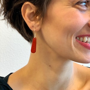 May include: A person wearing a pair of red, rectangular drop earrings. The earrings are a solid, deep red colour and hang from silver hooks. The person is smiling, and the background is a plain white wall.