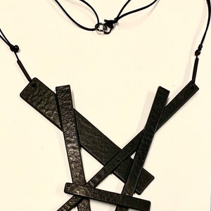 May include: Black leather necklace with an abstract design. The necklace features several rectangular leather pieces arranged in a layered, overlapping pattern. The necklace is suspended from a black cord with an adjustable clasp.