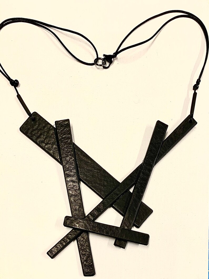 May include: Black leather necklace with an abstract design. The necklace features several rectangular leather pieces arranged in a layered, overlapping pattern. The necklace is suspended from a black cord with an adjustable clasp.